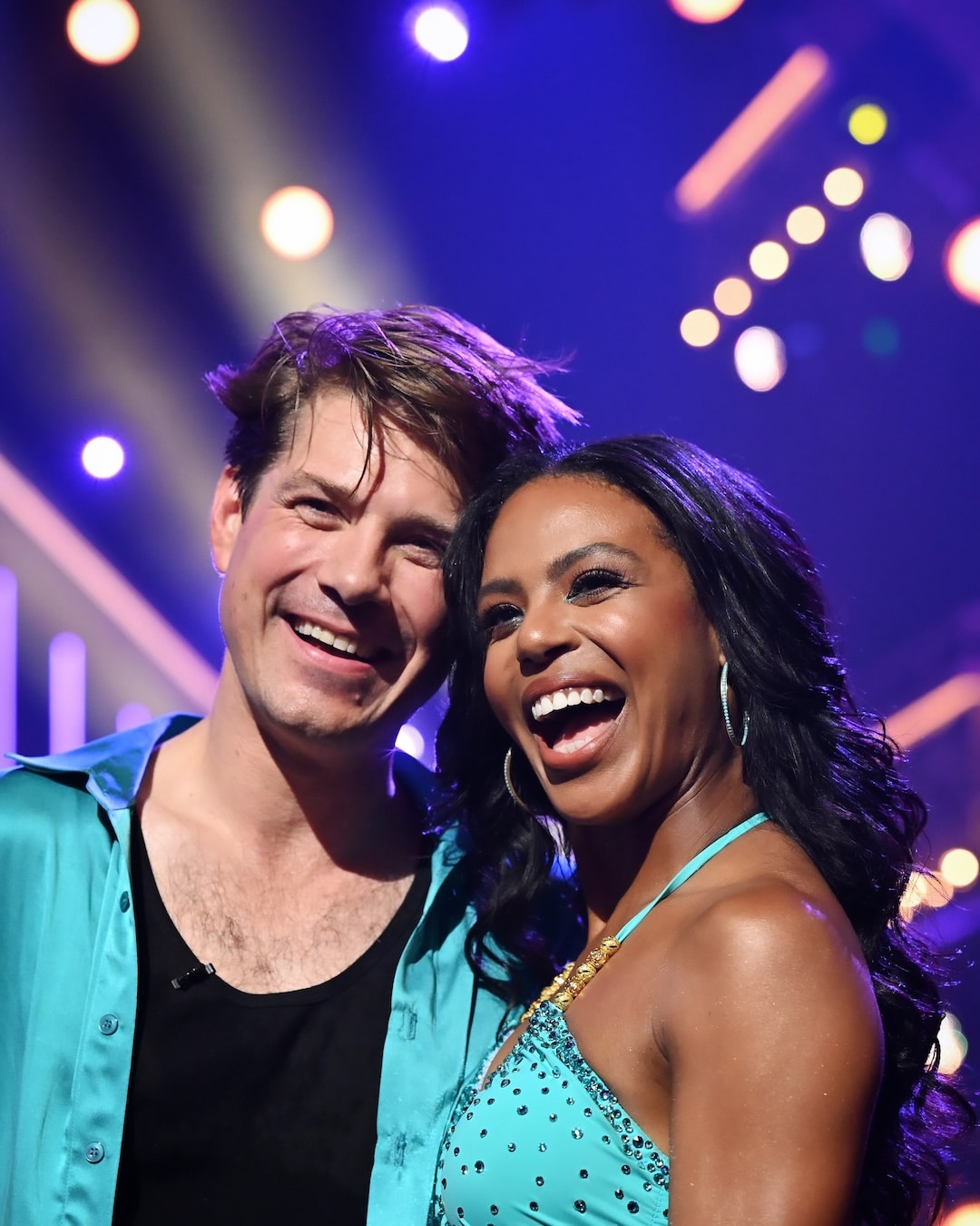 A smiling man and woman in matching teal dance outfits stand closely together on a brightly lit stage with colorful lights in the background, capturing a memorable moment from the Week 2 recap of Dancing With The Stars.