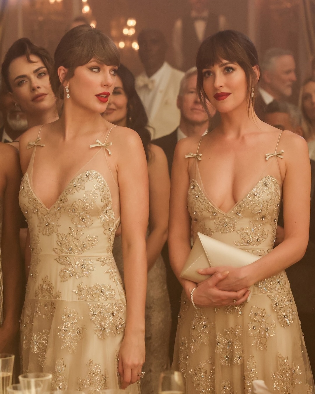Two women in matching gold, embellished dresses stand side by side at a formal event, surrounded by elegantly dressed guests in a softly lit room.