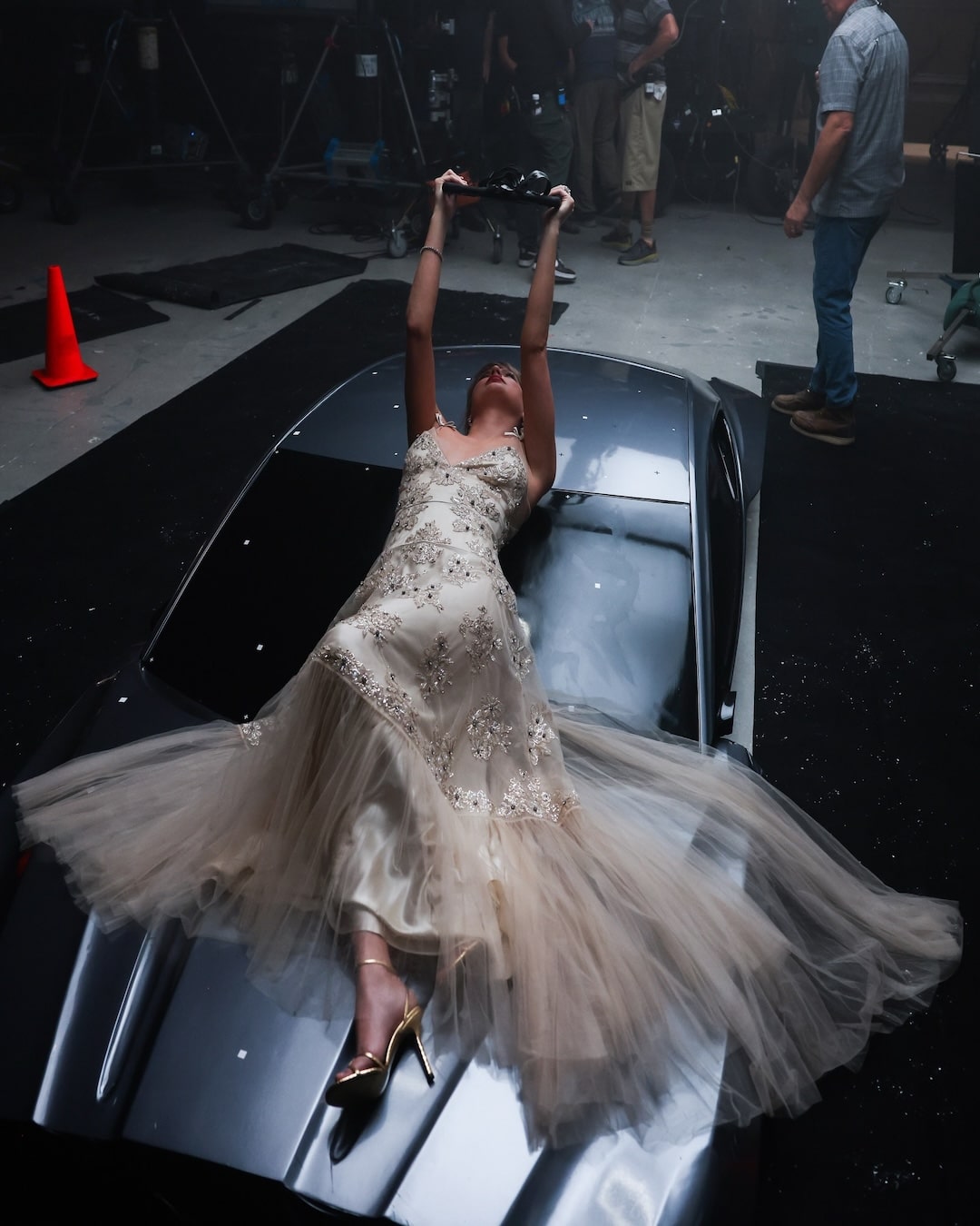 A woman in a long, elegant dress lies on the hood of a car, holding something above her head, surrounded by studio equipment and people in the background.