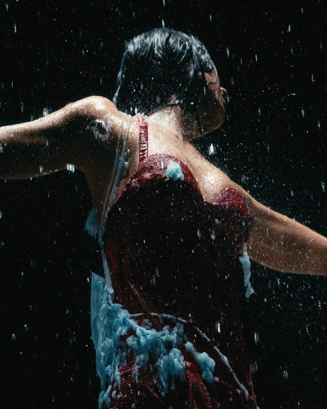 A person in a red dress stands with arms outstretched under falling water, with white foam on their dress and body, against a dark background.
