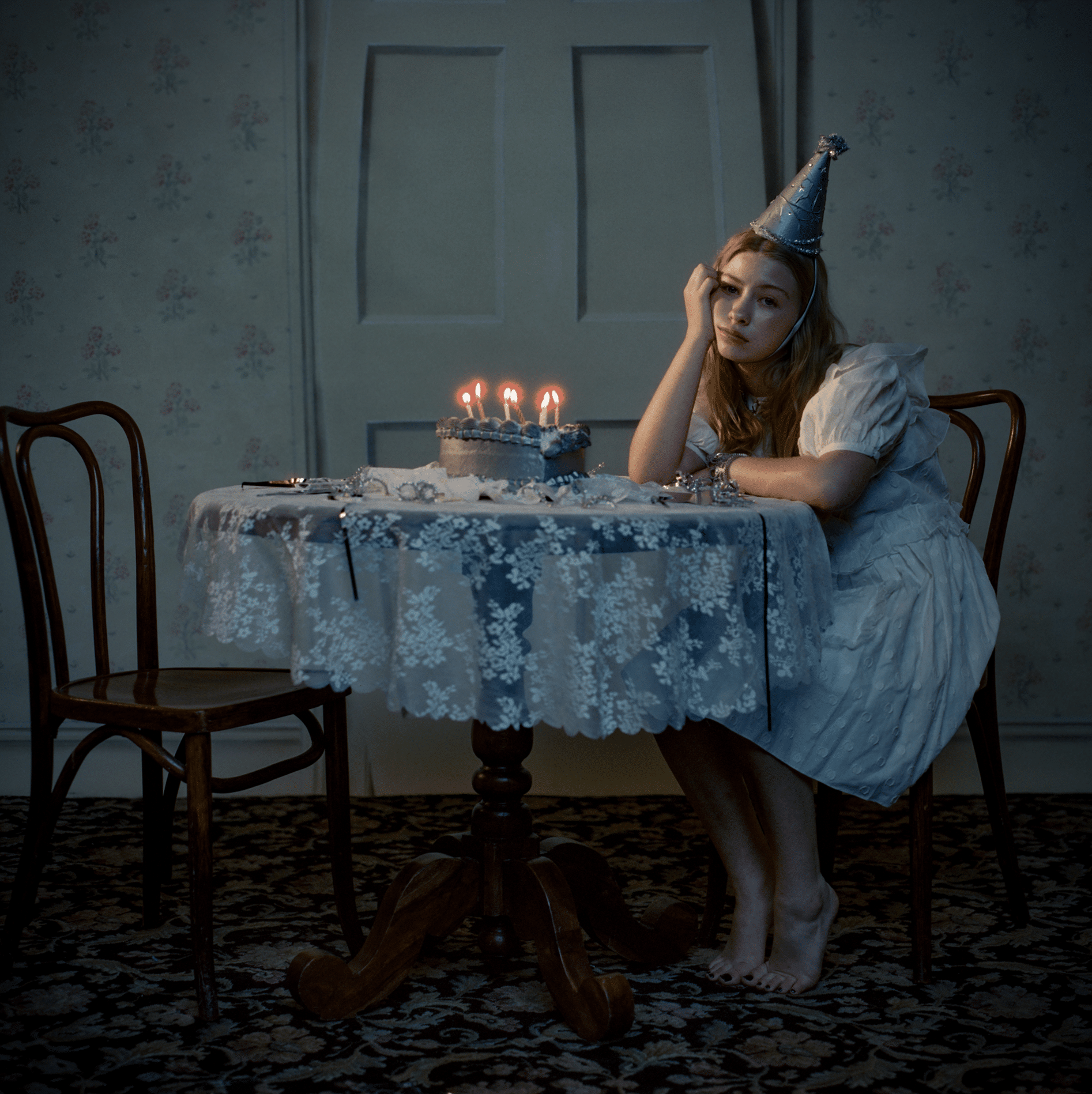 A sad young woman in a white dress and silver party hat sits alone at a round table with a lit birthday cake, resting her head on her hand. The dimly lit room, adorned with floral wallpaper and an empty chair, evokes the lonely, melancholic atmosphere of Blythe Woods.