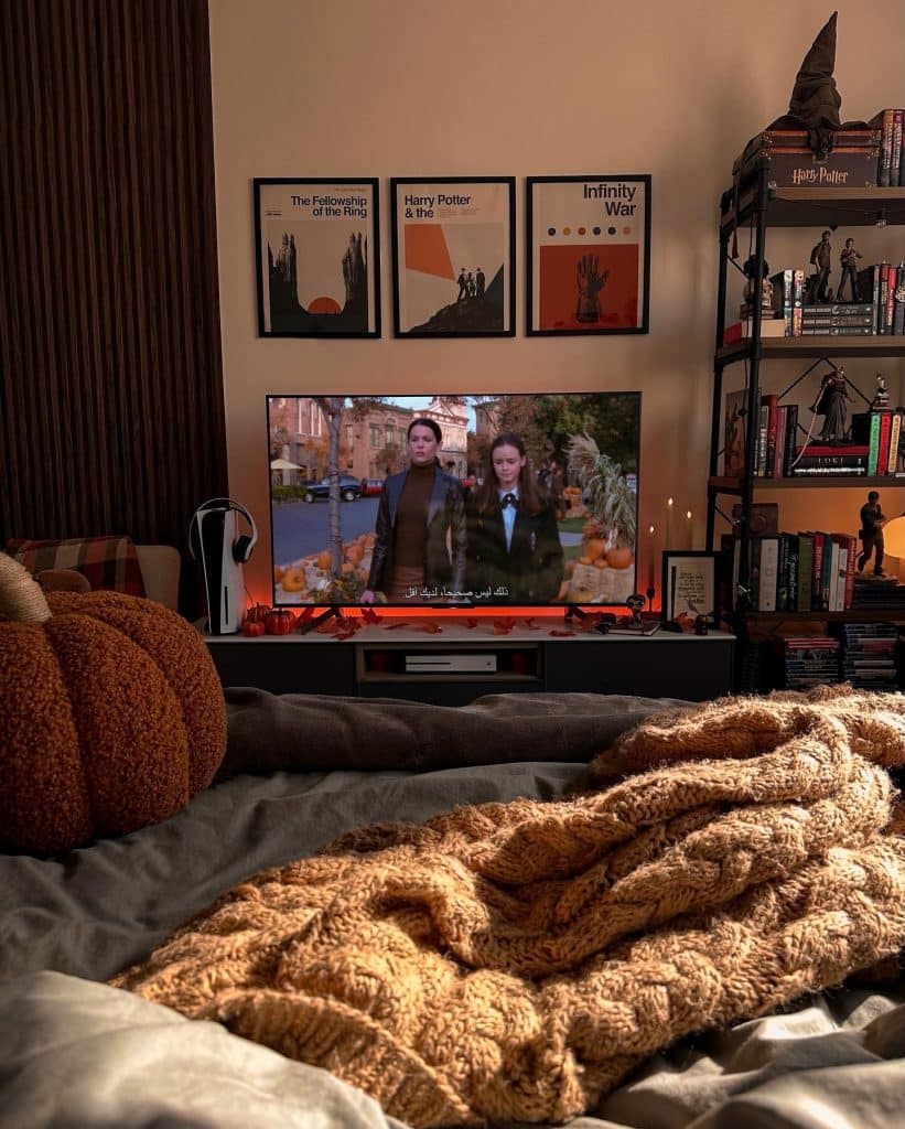 A cozy bedroom scene with a bed covered in knit blankets, a TV showing two people walking outdoors—perfect for your after-school routine or to binge-watch your favorite fall show. Shelves and wall art feature Harry Potter and Marvel themes, while warm lighting creates a relaxed atmosphere.