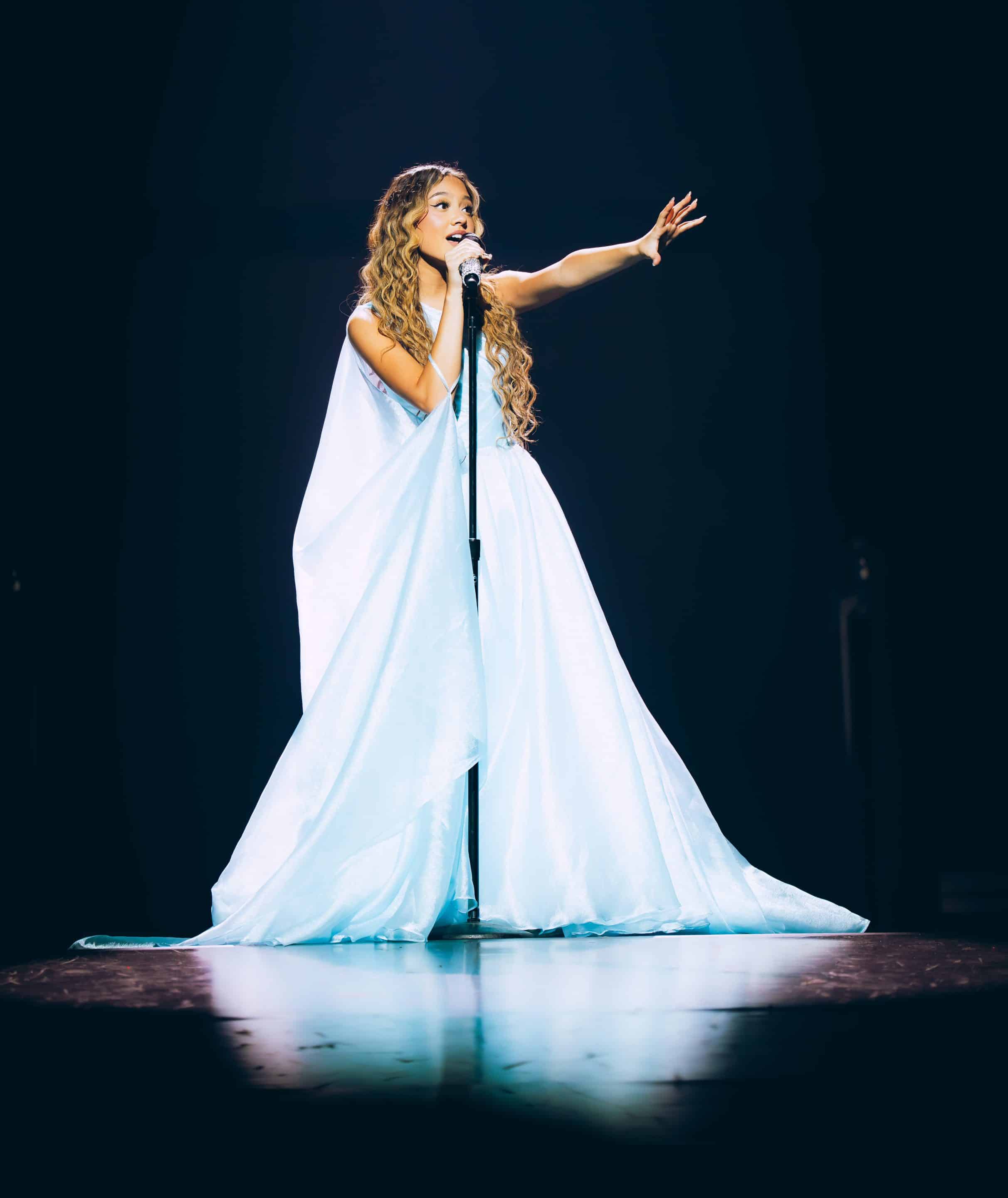 A woman in a flowing, light blue gown sings on stage, holding a microphone and reaching out with one hand—liamani segura spotlighted against a dark background.
