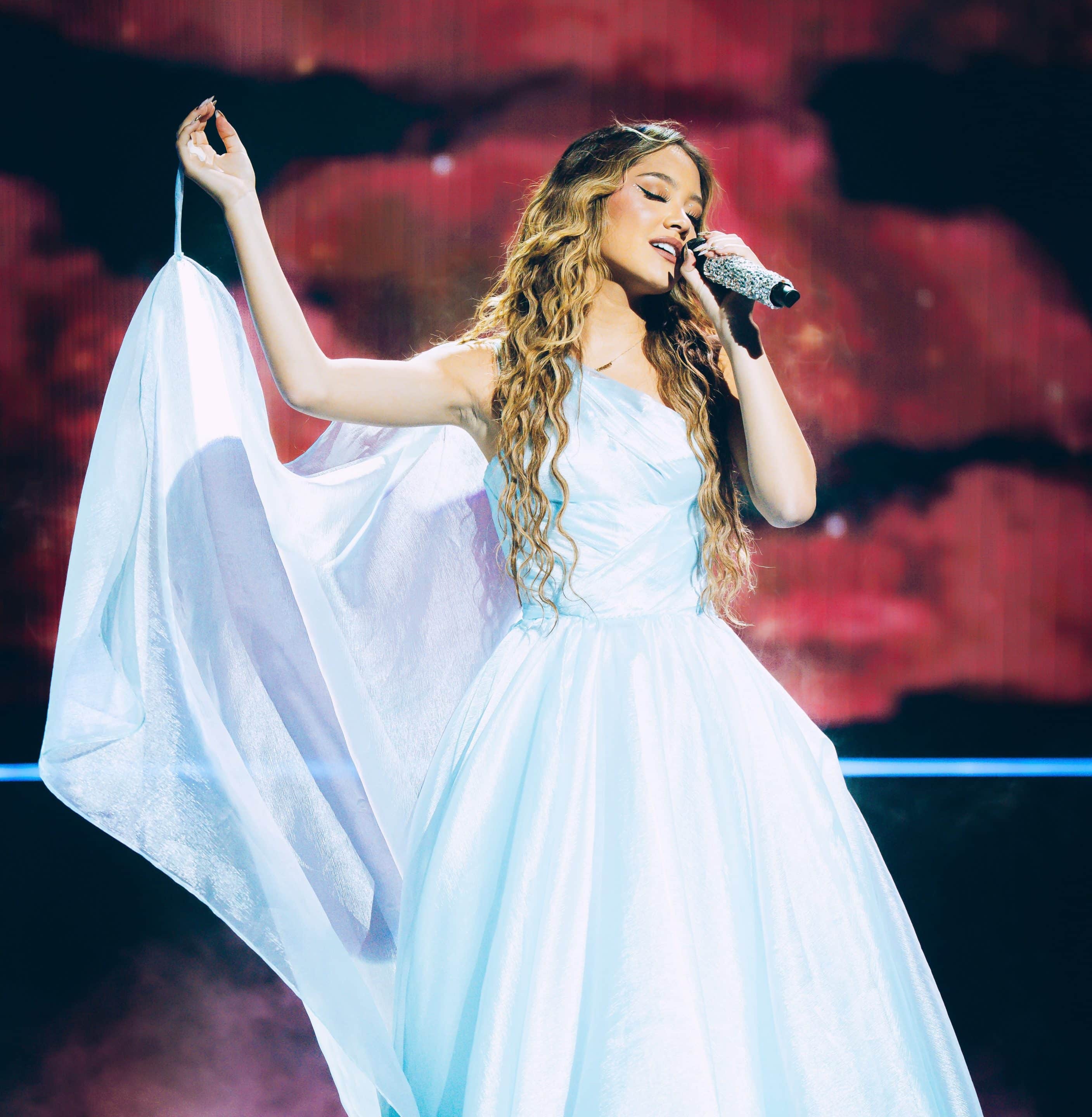 Liamani Segura, in a flowing white gown, sings passionately on stage, holding a microphone in one hand and lifting her dress with the other. A dramatic, colorful background enhances the performance ambiance.