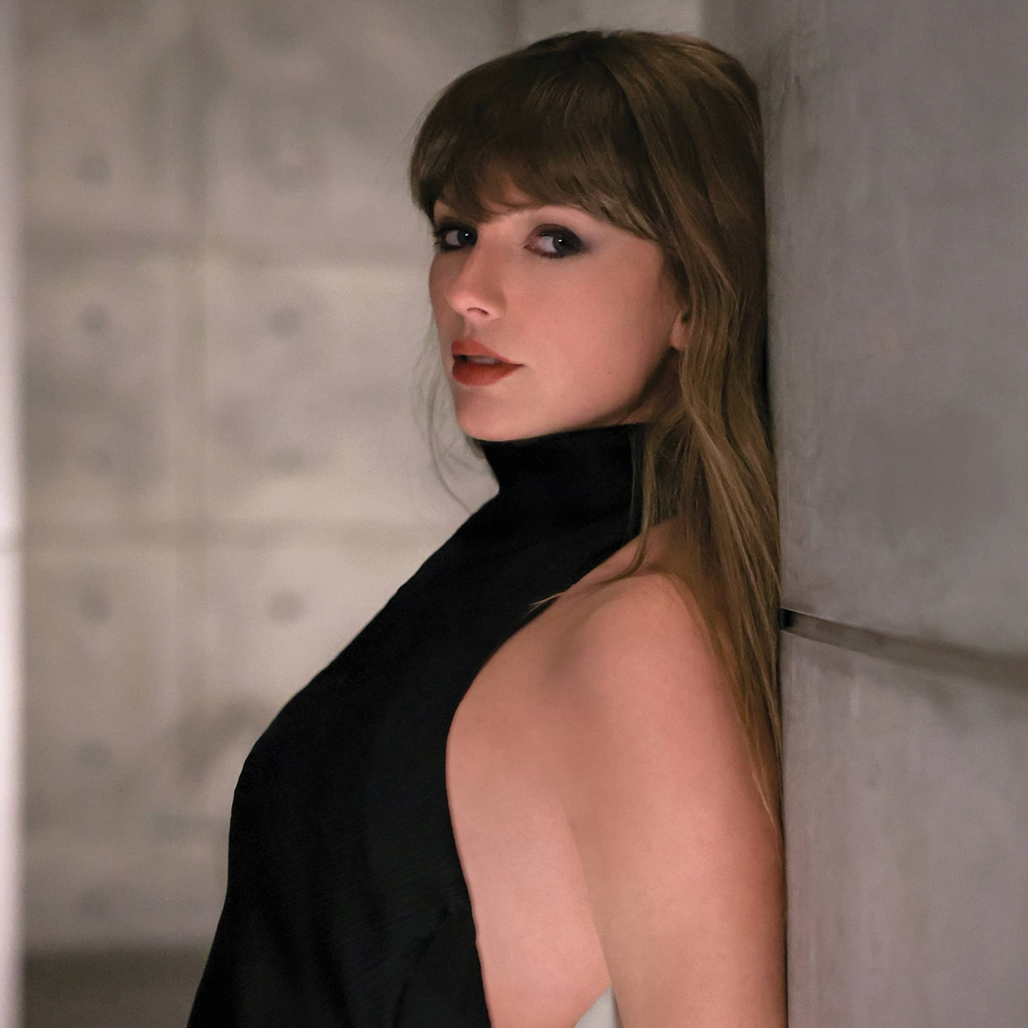 A woman with long brown hair and bangs, wearing a sleeveless black high-neck top, leans against a gray wall and looks toward the camera with a neutral expression, evoking the quiet confidence found in the life of a showgirl.