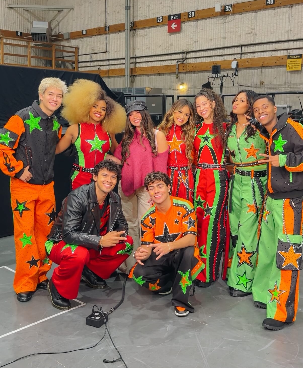 Nine people pose together indoors; seven wear matching green, orange, and black star-themed outfits, one wears a leather jacket, and one sports a casual pink top and hat. Smiling brightly, they appear to be part of a group—possibly performers or fans coming together in celebration at the Worlds Collide Concert Tour.